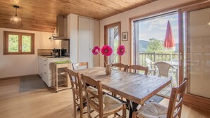 A wooden interior with a dining table, chairs, a kitchen area, and large windows offering a view of the outdoors.