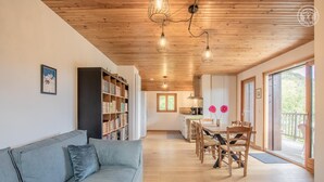 A spacious living area with a sofa, a dining table with chairs, a bookshelf, and a kitchen in the background.