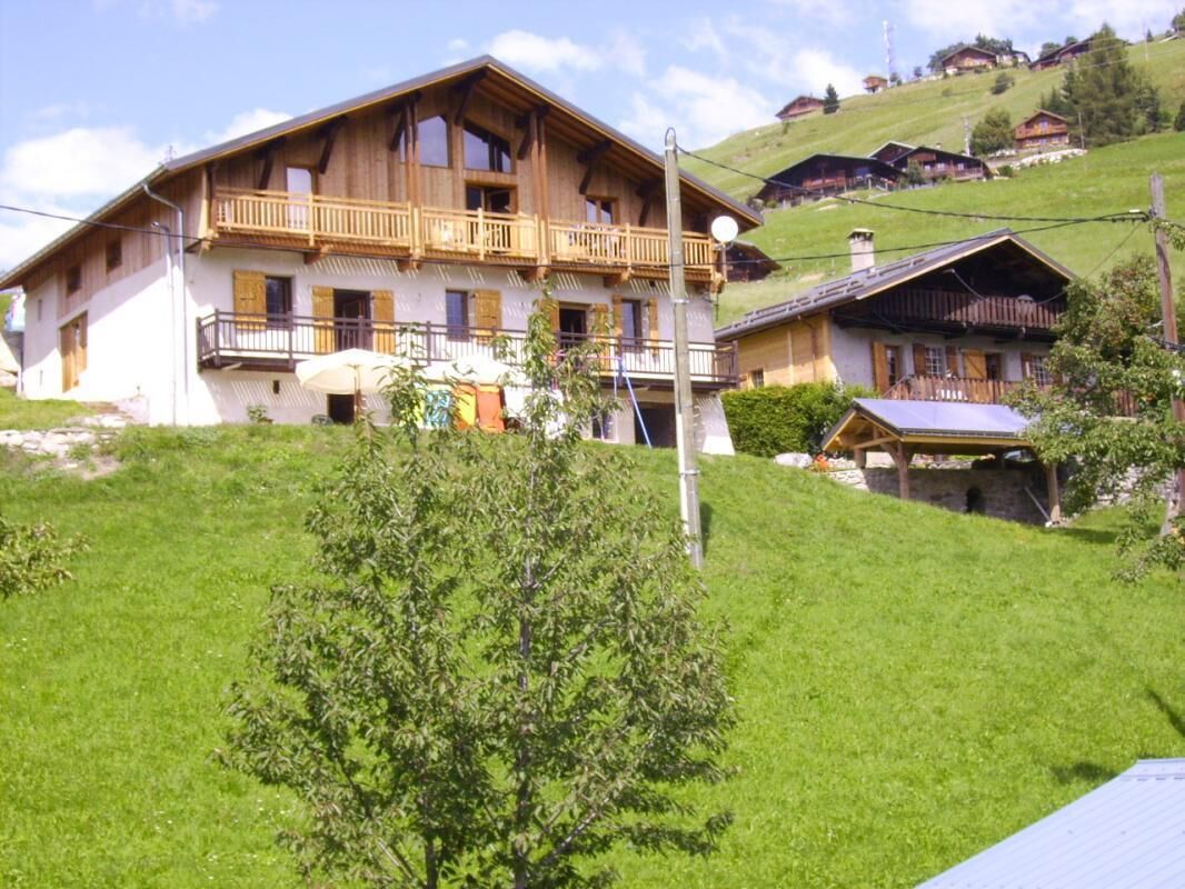 A large wooden chalet with a balcony, surrounded by lush green hills and other similar buildings.