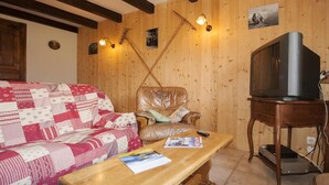 Ein gemütliches Wohnzimmer mit einer Holzwand, einem Ledersessel, einem Sofa mit Patchworkdecke, einem Holztisch mit Zeitschriften und einem Fernseher auf einem Holzständer.