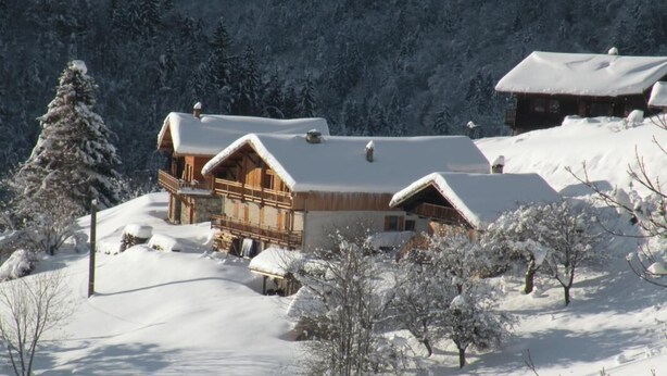 Eine Holzhütte mit schneebedecktem Dach, umgeben von schneebedeckten Bäumen und einer Bergkulisse.
