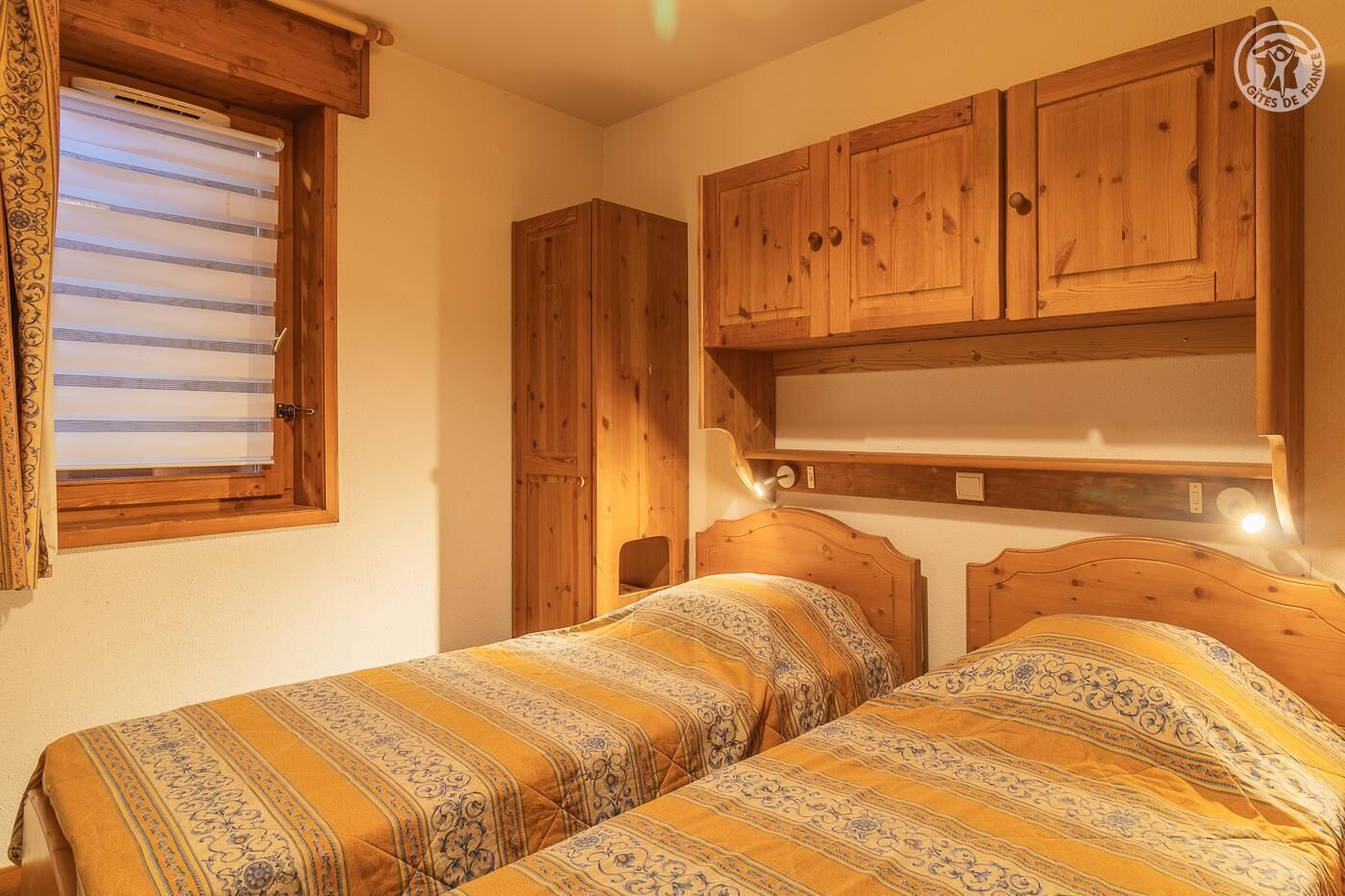 Quarto com duas camas, armários de madeira, janela com persianas e luminária sobre as camas.