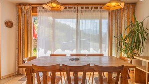 Un comedor con mesa y sillas de madera, ventanas amplias con cortinas traslúcidas y una planta en maceta.