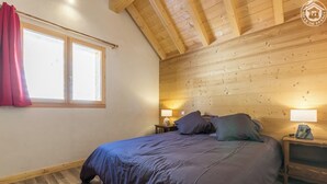 Un dormitori amb sostre de fusta, un llit gran amb roba de llit fosca, dues làmpades de nit i una finestra amb cortines.