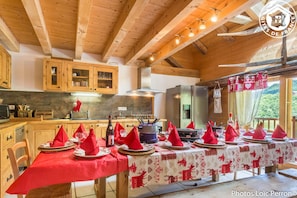 Comedor con mesas de madera dispuestas para una comida, cocina al fondo y vista a zonas verdes exteriores.