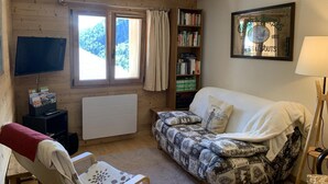 A cozy room with a wooden ceiling, a sofa, a bookshelf, a television, and a window with curtains. - Chalet LE Char - Gite "LE Plan" (LA GIETTAZ)