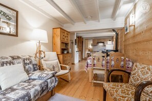 A cozy living room with a sofa, armchair, and wooden dining table. A kitchen is visible in the background.