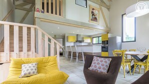 Un vaste espace de vie avec un canapé jaune et un fauteuil marron, une table à manger avec des chaises jaunes, et une cuisine en arrière-plan.