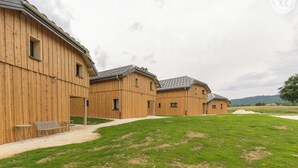 Une rangée de granges en bois, reliées par un chemin de gravier, se dresse au milieu d’un champ verdoyant, sous un ciel nuageux.