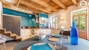A modern kitchen with wooden beams, a dining area with a table set for two, and a large window with a view of greenery. - Clos DES Platanes-cote Figuier (BRISON SAINT INNOCENT)