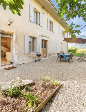 Un bâtiment rustique en pierre, doté d’une cour en gravier, d’un espace extérieur aménagé pour s’asseoir et d’un barbecue.