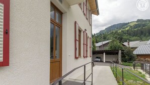 A building with a white exterior, red shutters, and a wooden door. There is a concrete pathway leading to a metal railing. - DE Saint Jean Baptiste (BEAUFORT ARECHES)