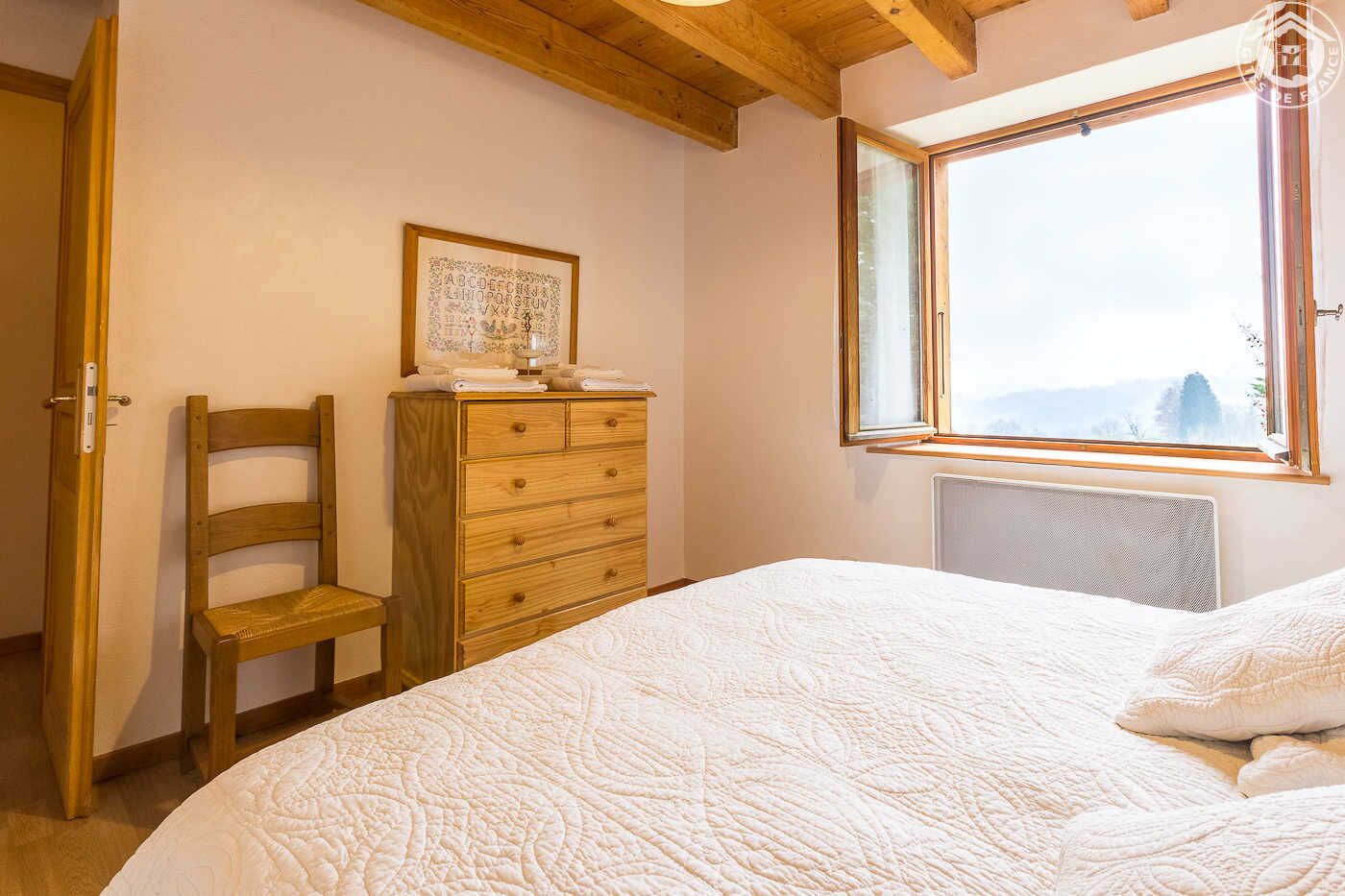 Ein Schlafzimmer mit einem Holzkasten, einem Sessel, einem Bett mit weißer Bettdecke und einem Fenster mit Blick ins Freie.