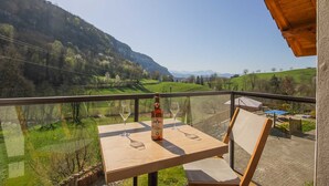 Une table en bois, sur laquelle se trouvent une bouteille et un verre, est placée sur un balcon donnant sur une vallée verdoyante et des montagnes.