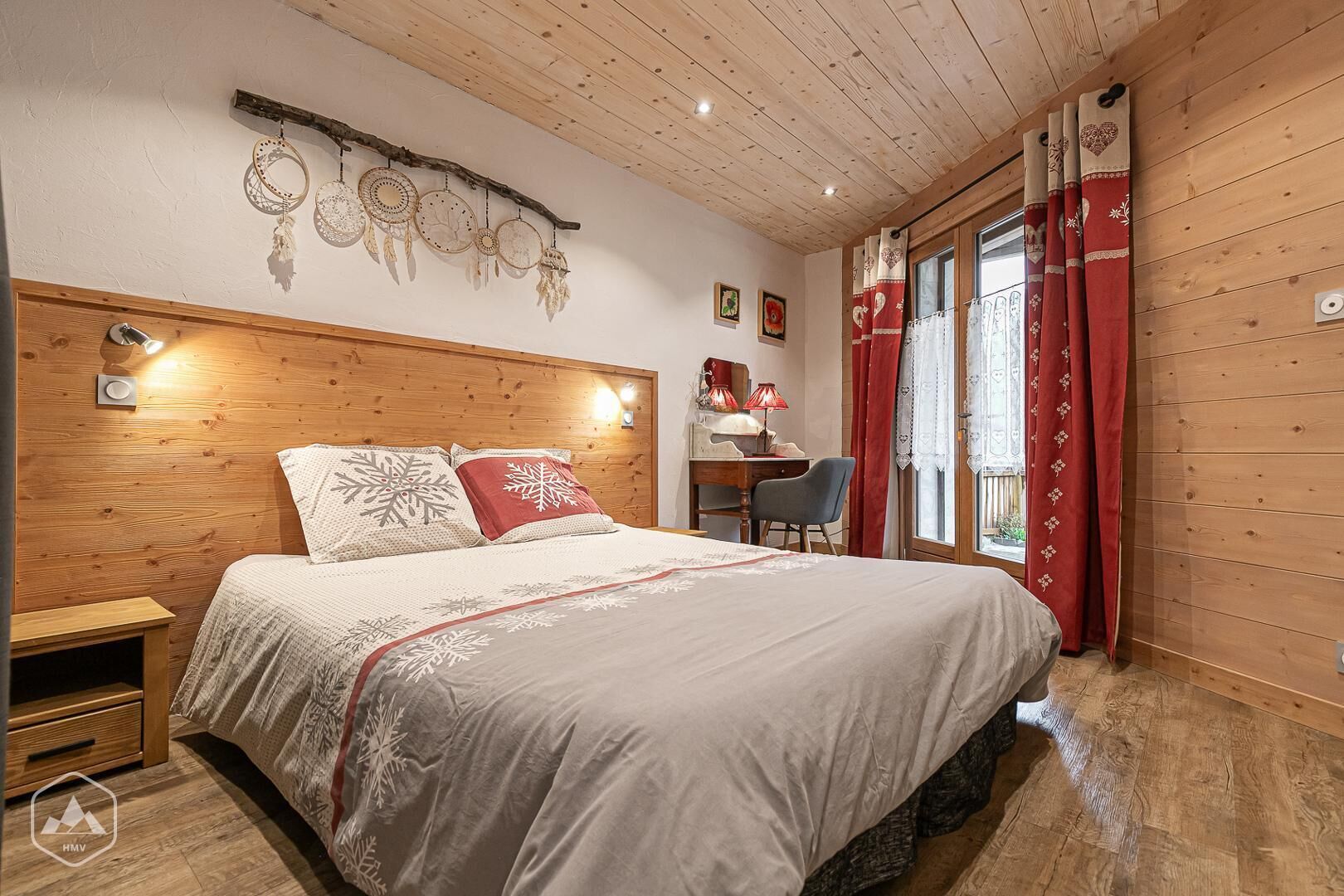 Een knus slaapkamer met een houten hoofdbord, een bed met sneeuwvlindes op het beddengoed, een bureau met een lamp en een raam met rode gordijnen.