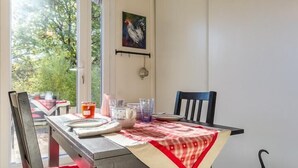 Un coin repas avec une table dressée pour deux personnes, des chaises, un luminaire suspendu et une vue sur la verdure à l’extérieur.