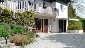 Une maison à deux étages avec un balcon, une allée gravillonnée et un jardin agrémenté de fleurs et d’arbustes.
