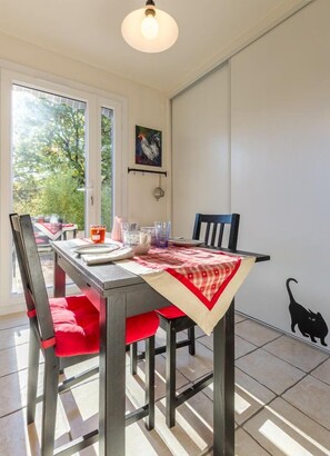 Een eethoek met een tafel voor twee personen, stoelen, een hanglamp en uitzicht op groen.