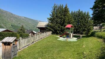 Un terrain de jeux avec un toboggan et des balançoires, entouré d’une clôture en bois, avec en arrière-plan des maisons et des montagnes.