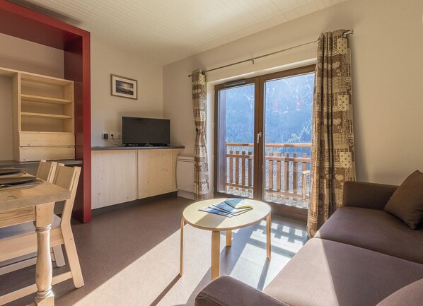 A living room with a sofa, a dining table with chairs, a television on a wooden cabinet, and a balcony with patterned curtains.