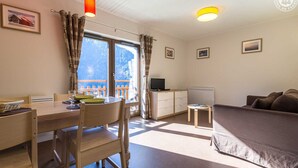 A room with a wooden dining table, chairs, a small TV, a sofa, and a window with curtains. - LA Lauziere - Montsapey (MONTSAPEY)
