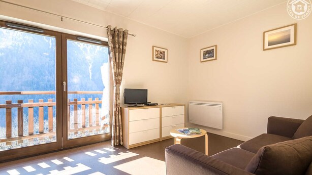 A living room with a sofa, a TV on a wooden cabinet, a radiator, and a window with a view of a wooden deck. - LA Lauziere - Montsapey (MONTSAPEY)