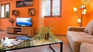 A living room with a television, a glass coffee table with magazines, a sofa, and a potted plant.