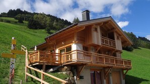 A wooden chalet with a balcony, situated on a hillside with a signpost indicating nearby locations.