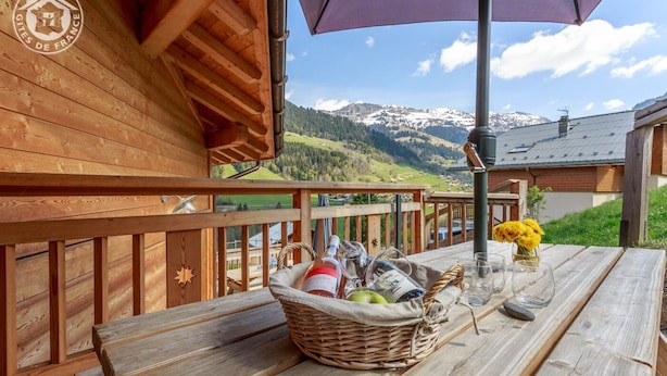 A wooden deck with a view of snow-capped mountains, a wicker basket with bottles, and a vase with yellow flowers.