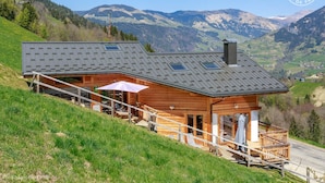 A wooden chalet with a sloped roof, a balcony, and a view of the surrounding mountains.