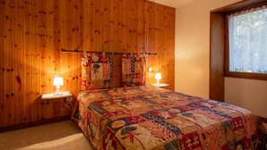 Ein Schlafzimmer mit Holzwandverkleidung, einem Bett mit bunter Steppdecke, zwei Nachttischlampen und einem Fenster mit Spitzenvorhängen.