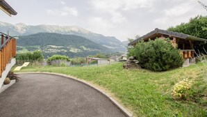 A rural property with a paved driveway leading to a wooden cabin, surrounded by lush greenery and mountains in the background.