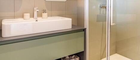 A modern bathroom with a glass-enclosed shower, a white sink with a mirror above it, and a shelf below the sink.