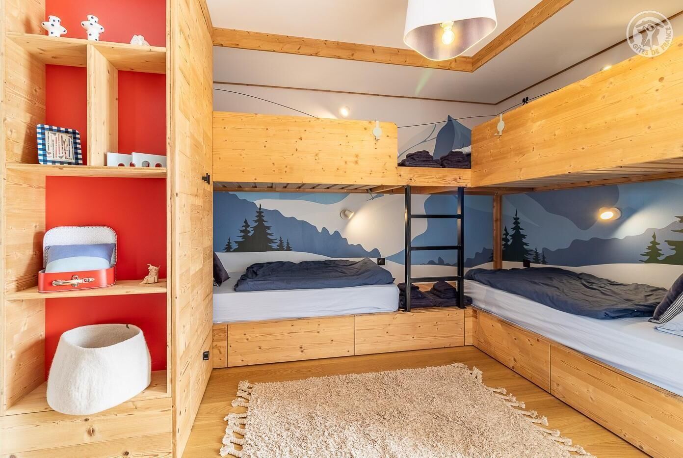 A cozy room with two bunk beds, a wooden shelf with decorative items, a white cat bed, and a mountain-themed mural on the wall.