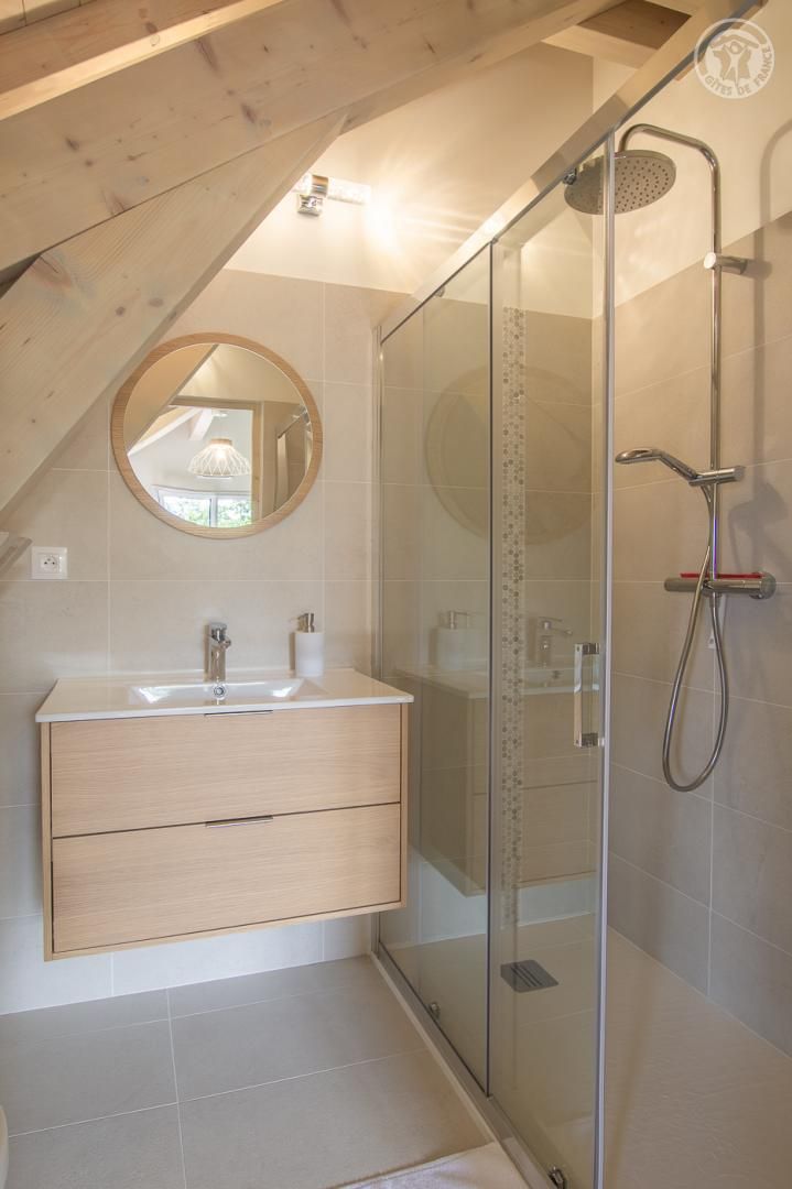 A modern bathroom with a glass shower enclosure, a wooden vanity with a round mirror, and a tiled floor.