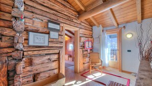 A log cabin interior with wooden walls, a wooden bench, a wicker chair, and a door leading to another room. - Chalet Lidia Ski in-Ski out 50m (Livigno)