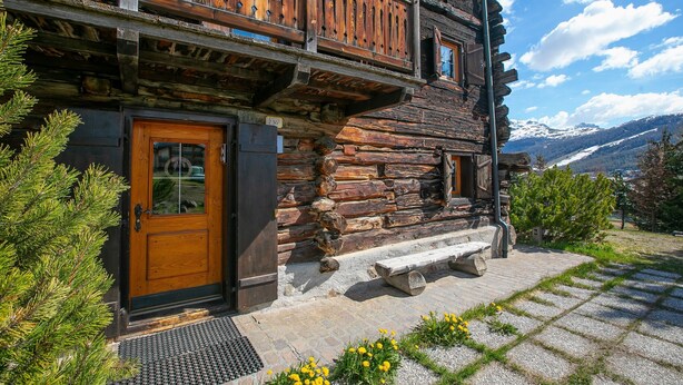 A log cabin with a wooden door, a stone bench, and a view of snow-capped mountains. - Chalet Lidia Ski in-Ski out 50m (Livigno)