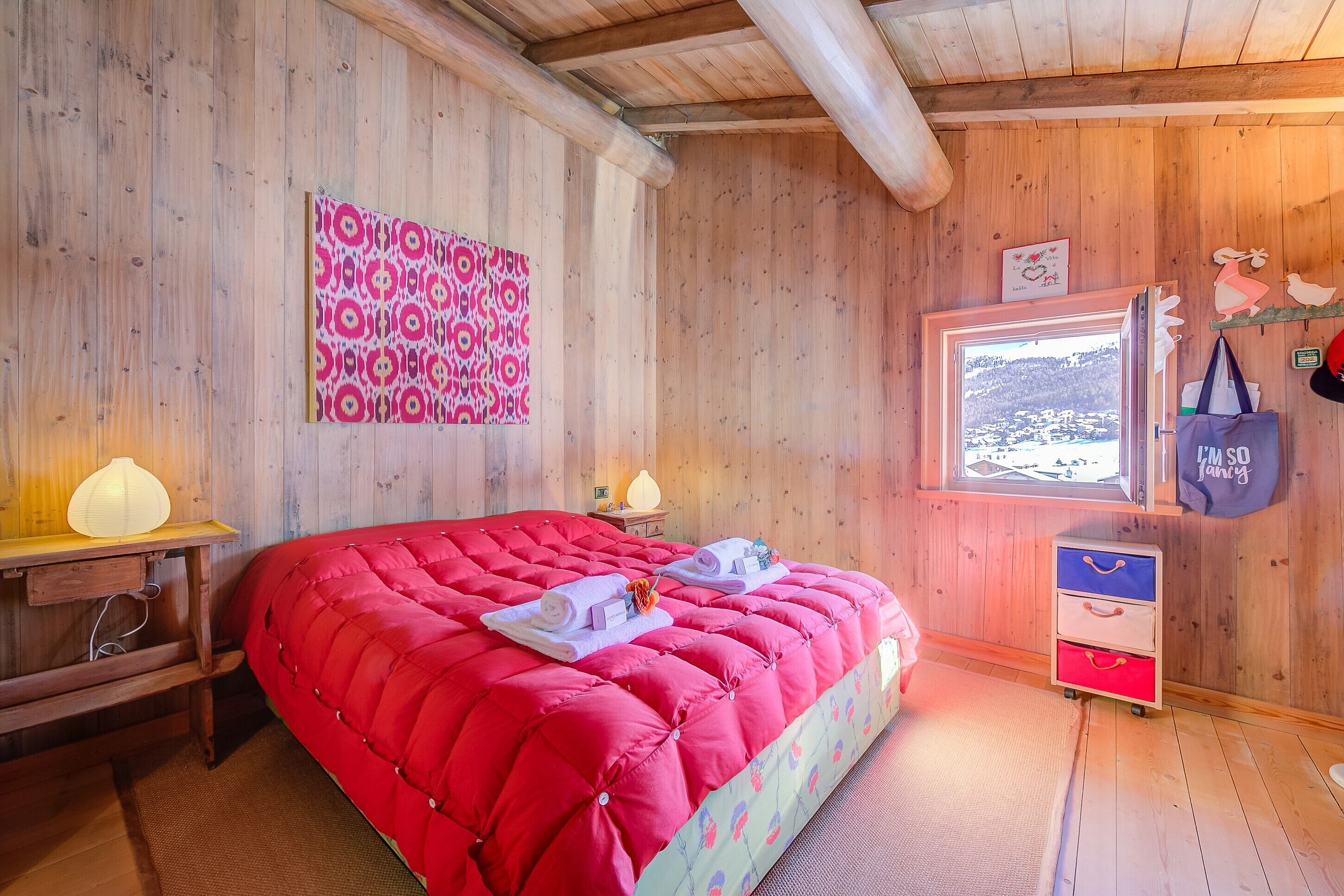 A wooden room with a bed, a desk, a lamp, a window with a view, and a wall decoration.