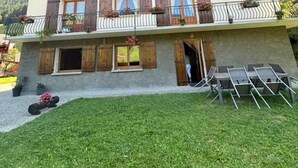Un chalet de madera de dos pisos con balcón y un pequeño espacio exterior para sentarse, con montañas al fondo.
