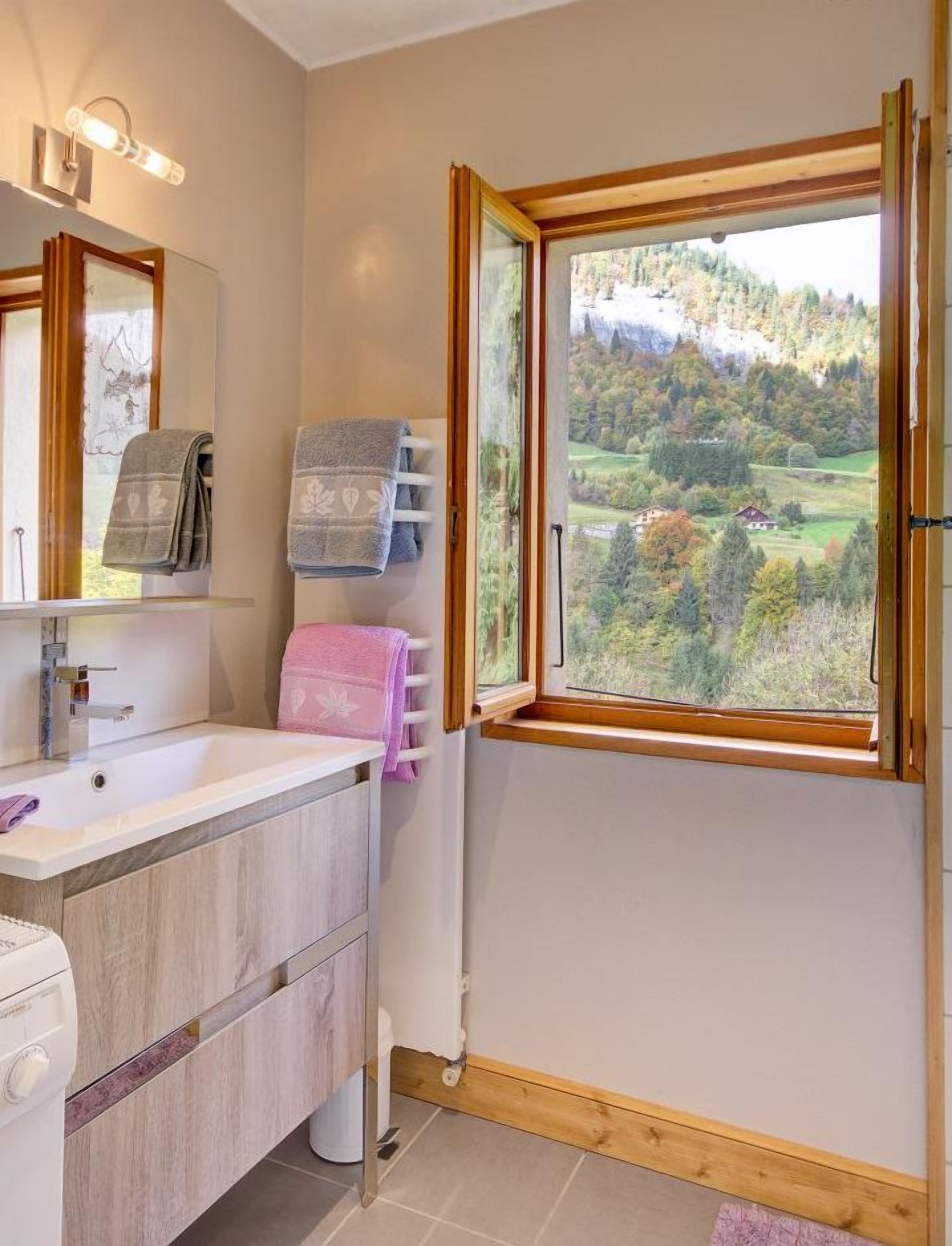 Un bagno con un mobile bagno in legno, uno specchio, una finestra vista su un paesaggio boschivo e un portaservizi con diversi asciugamani.