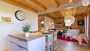 Cozinha acolhedora com ilha de madeira, sala de jantar com tapete vermelho e sala de estar com sofá e televisão.