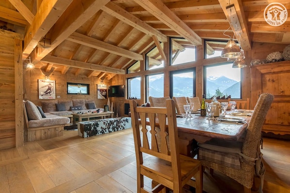 Cozy chalet interior with a dining area, comfortable sofa, and large windows offering a view of the surrounding landscape.