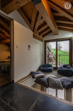 A room with a high ceiling, wooden beams, a sliding glass door leading outside, and a small seating area with bean bags and cushions.
