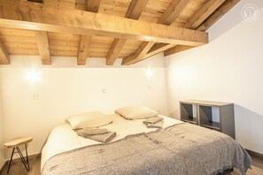 Quarto com teto de madeira, cama com almofadas e cobertor, um banquinho pequeno e um suporte para TV.