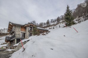 A chalet-style building with a stone facade, wooden balconies, and a ski slope in the background.