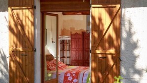 Una habitación rústica con una cama, contraventanas de madera y vistas a un portón rojo.