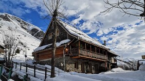 Un chalet traditionnel, avec une façade en bois, des fondations en pierre et un balcon donnant sur un paysage enneigé, avec des montagnes en arrière-plan.