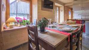Un intérieur en bois comprenant une table à manger pour quatre personnes, un téléviseur fixé au mur et un coin cuisine équipé d’appareils modernes.