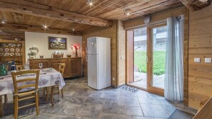 A wooden interior with a dining table set for two, a refrigerator, and a view through a glass door to an outdoor area.