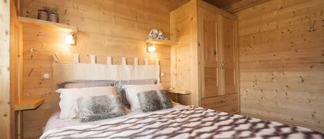 Un dormitorio de cabaña de madera con cama, almohadas, mesita de noche y estante con objetos decorativos.